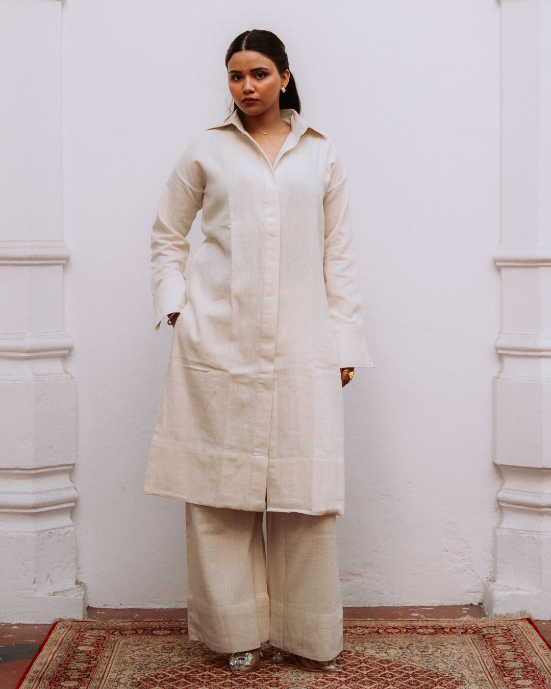 Woman wearing a long beige coat standing against a white wall. Sarmad Clothing collection by sukhanvar Mehr Co Ord Set Kurta Pant