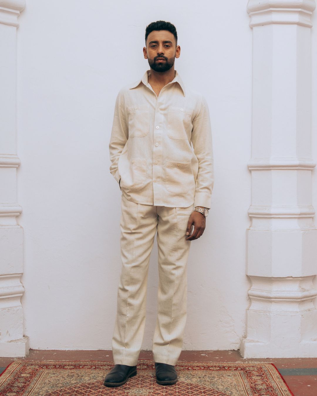 Man wearing a beige outfit standing against a white wall with decorative columns. Sarmad Clothing collection by sukhanvar Mens Raavi Co ord Set 4 Pocket Shirt and Trouser Kala Cotton from kutch Bhujodi