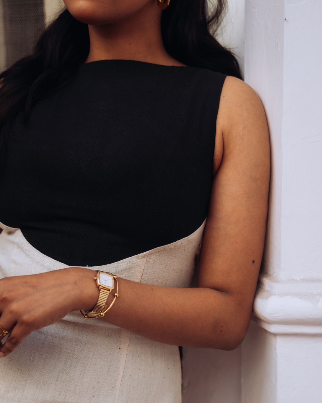 Mehvar dress Black on Ivory in Kala Cotton — Sukhanvar Sarmad SS2026. Woman wearing a black sleeveless top and gold watch, leaning against a white wall. 