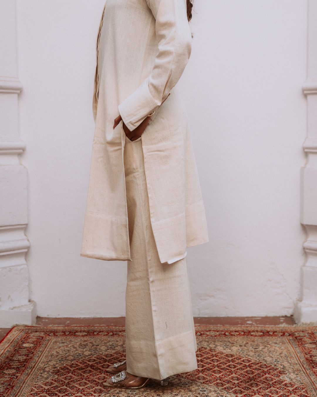 Person wearing a beige coat and pants standing on a patterned rug with a white wall background Sarmad Clothing collection by sukhanvar Mehr Co Ord Set Kurta Pant