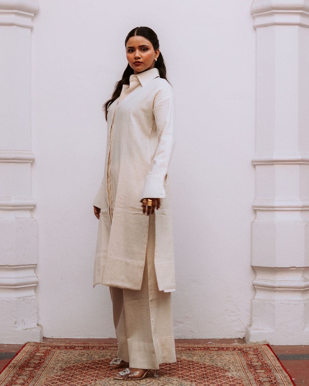 Woman wearing a beige coat and pants standing against a white wall. Sarmad Clothing collection by sukhanvar Mehr Co Ord Set Kurta Pant
