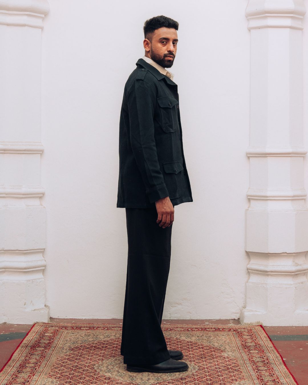 Man in a dark suit standing against a white wall with decorative columns. Sarmad Clothing collection by sukhanvar Mens Shacket jacket blazer Raahgir in Kala Cotton from kutch Bhujodi
