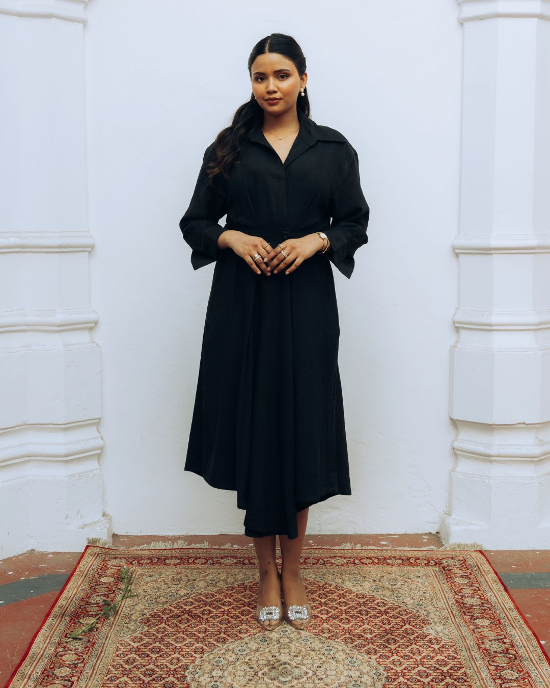 Woman wearing a black dress standing against a white wall with a patterned rug on the floor. Sarmad Clothing collection by sukhanvar Raqs Flowing Dress in KalaCotton from kutch Bhujodi