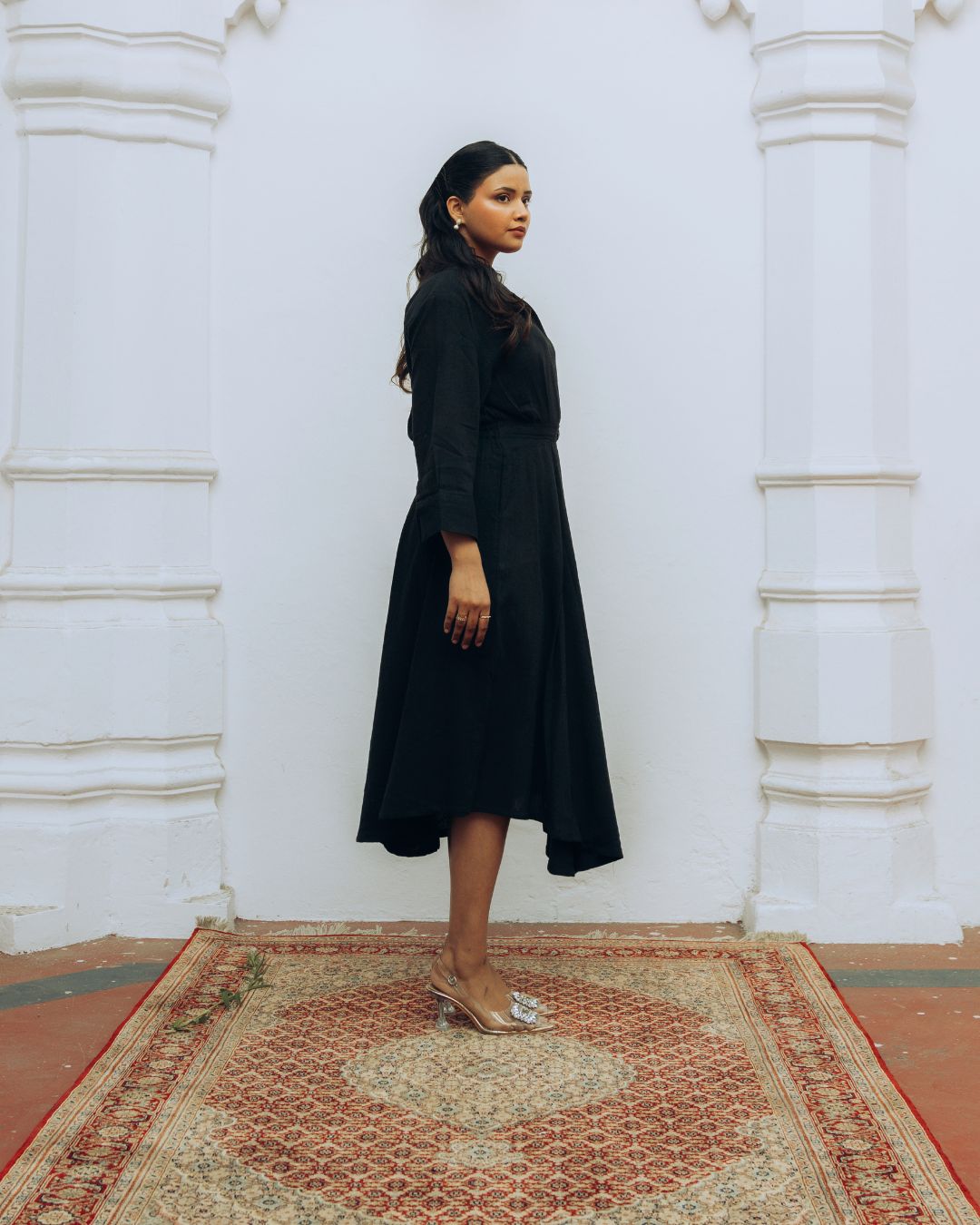 Woman in a black dress standing on a patterned rug with white walls in the background Sarmad Clothing collection by sukhanvar Raqs Flowing Dress in KalaCotton from kutch Bhujodi