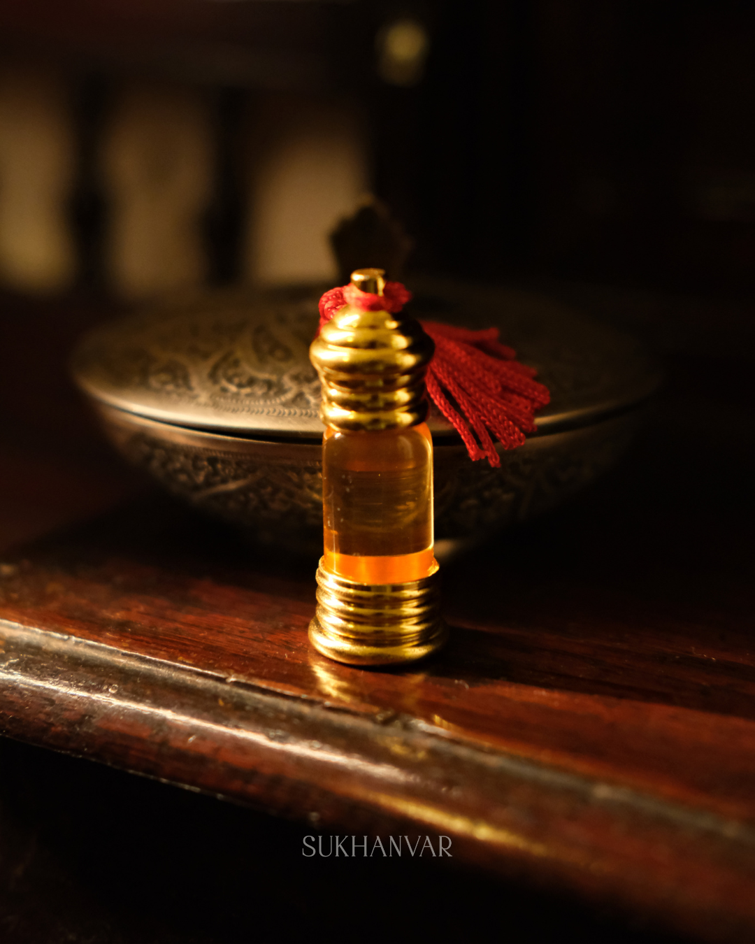 Sukhanvar Visaal Attar white musk perfume oil in a gold-capped glass bottle with a red tassel, set against a warm candlelit background.